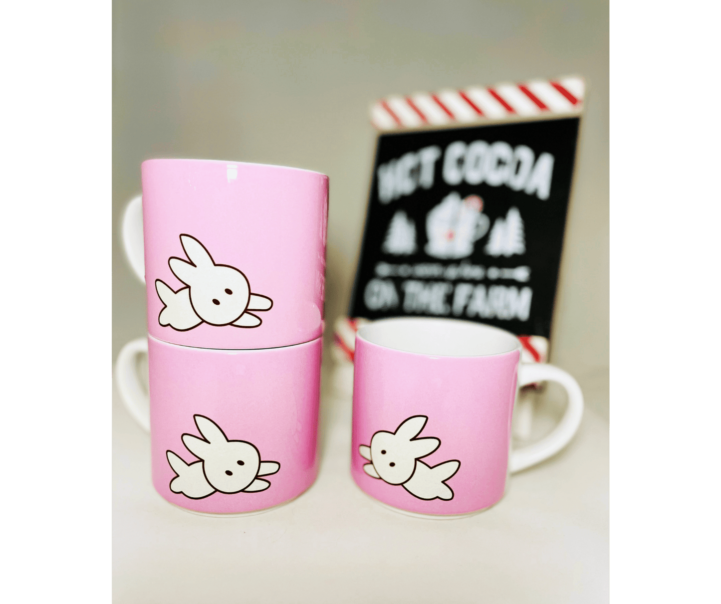 3 pink stackable kawaii bunny mugs. 10 oz mugs in front of a white background and hot coco sign