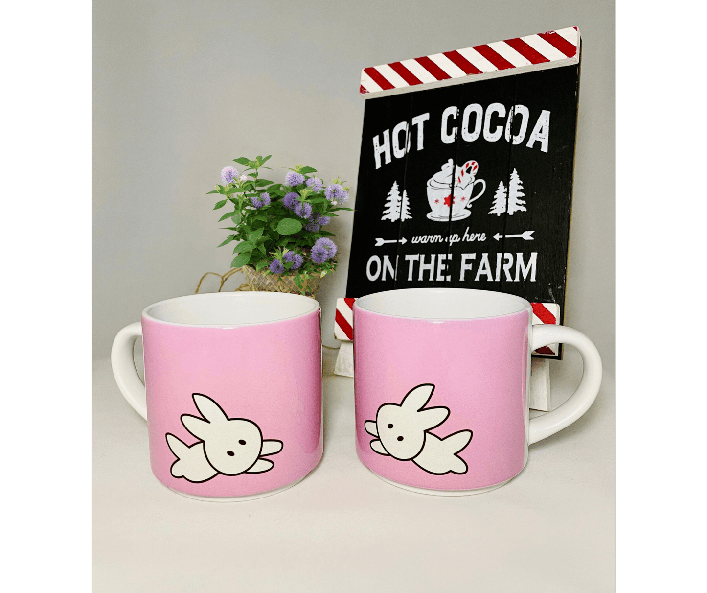 2 pink stackable 10 oz mugs in front of a white background and hot coco sign with a plant